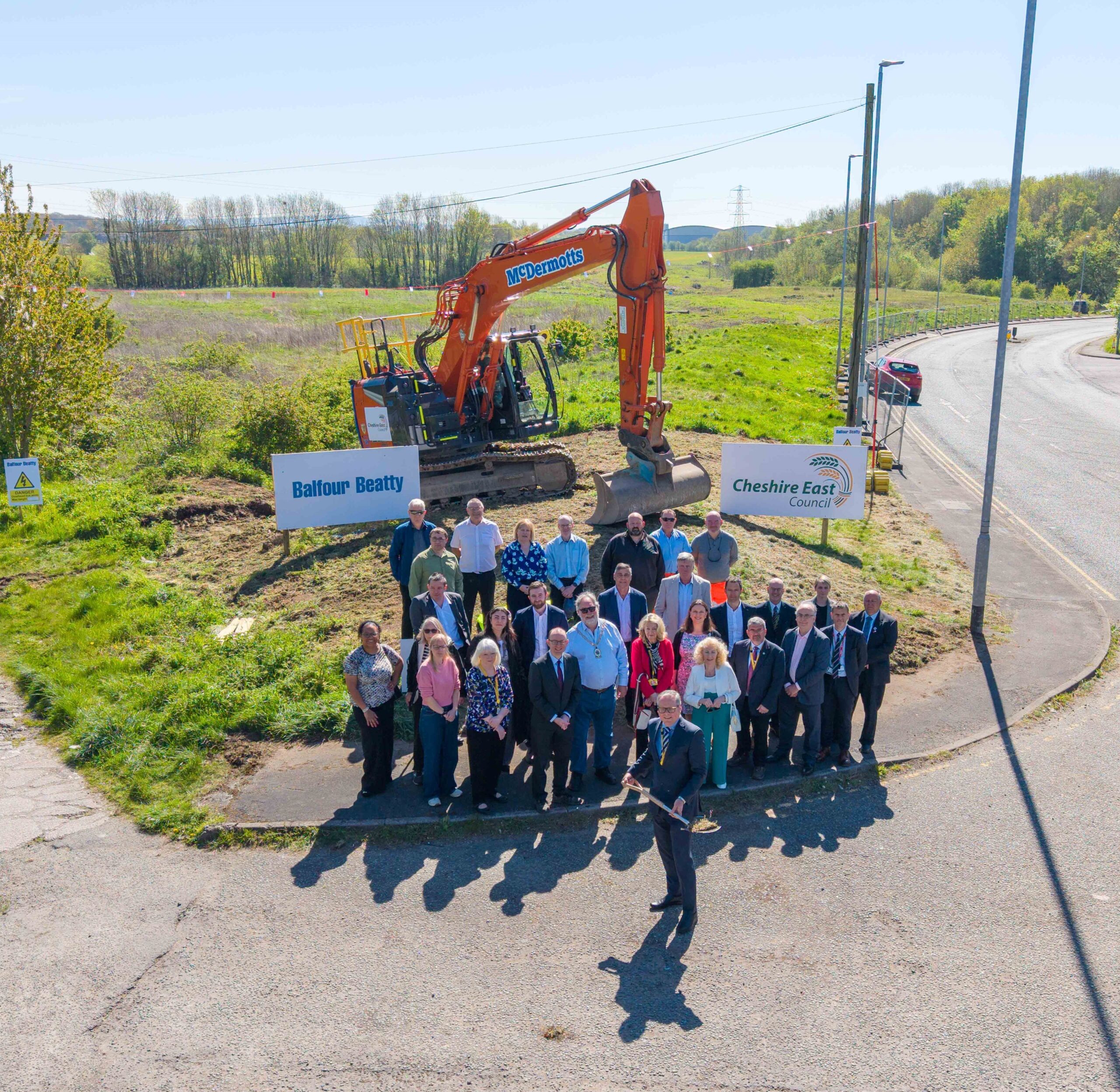 A group of Balfour Beatty members in attendance at sod cutting event for Middlewich Eastern Bypass, standing in front of a patch of grass on Bypass, in front of an orange digger