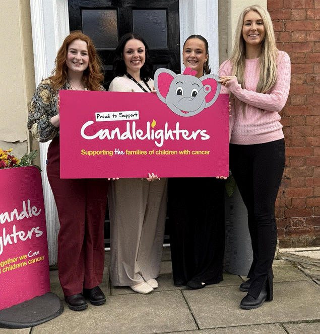 Four women stood outside the door of a small building, holding a large rectangular 'Candlelighters support' card