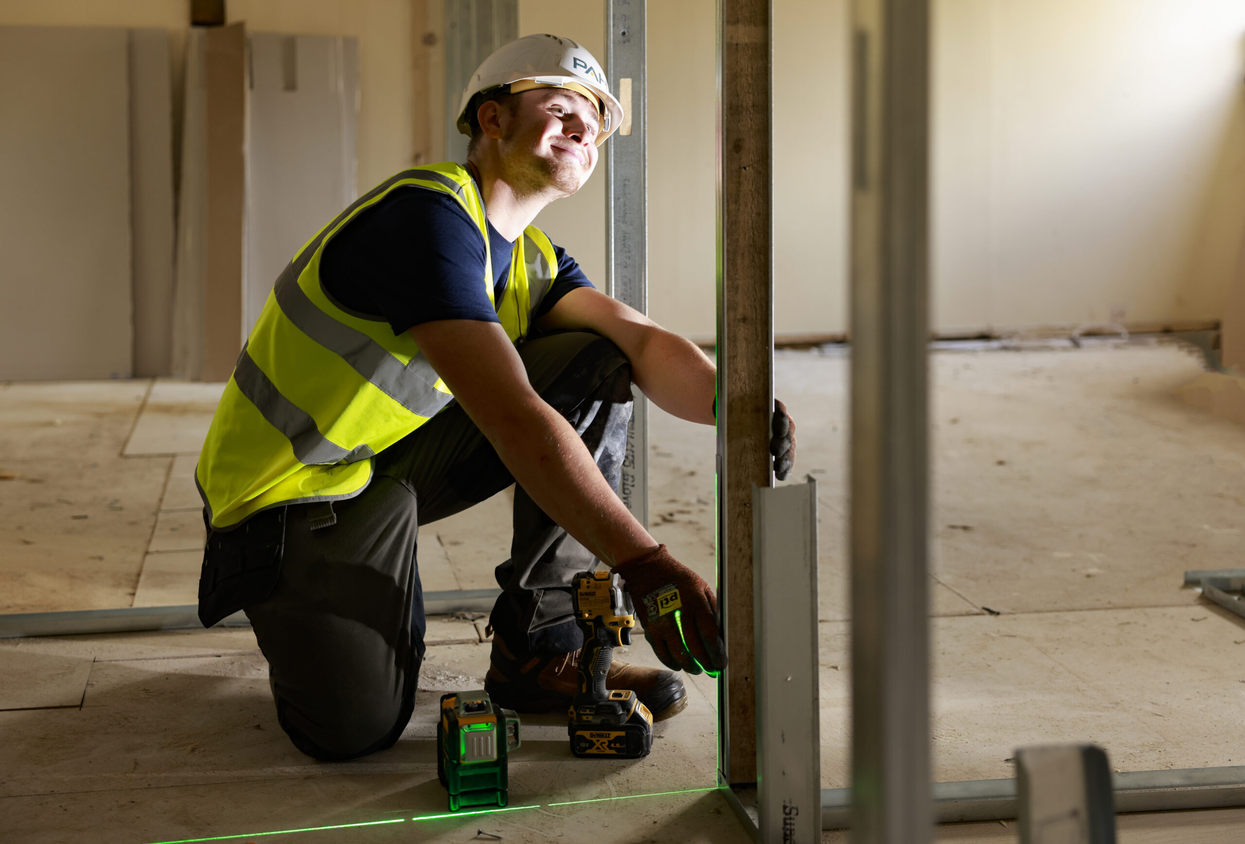 A PAH apprentice crouched down, wearing a high-vis and a hard white hat in a building that is being built
