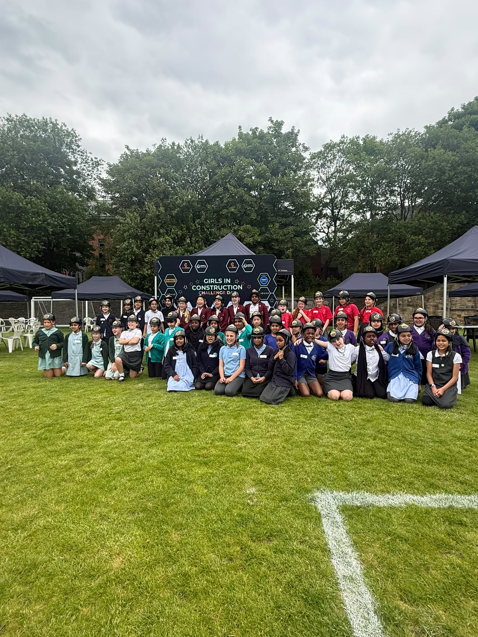 50 primary school girls wearing black building hats, in front of the 'Girls in Construction' sign on the school field