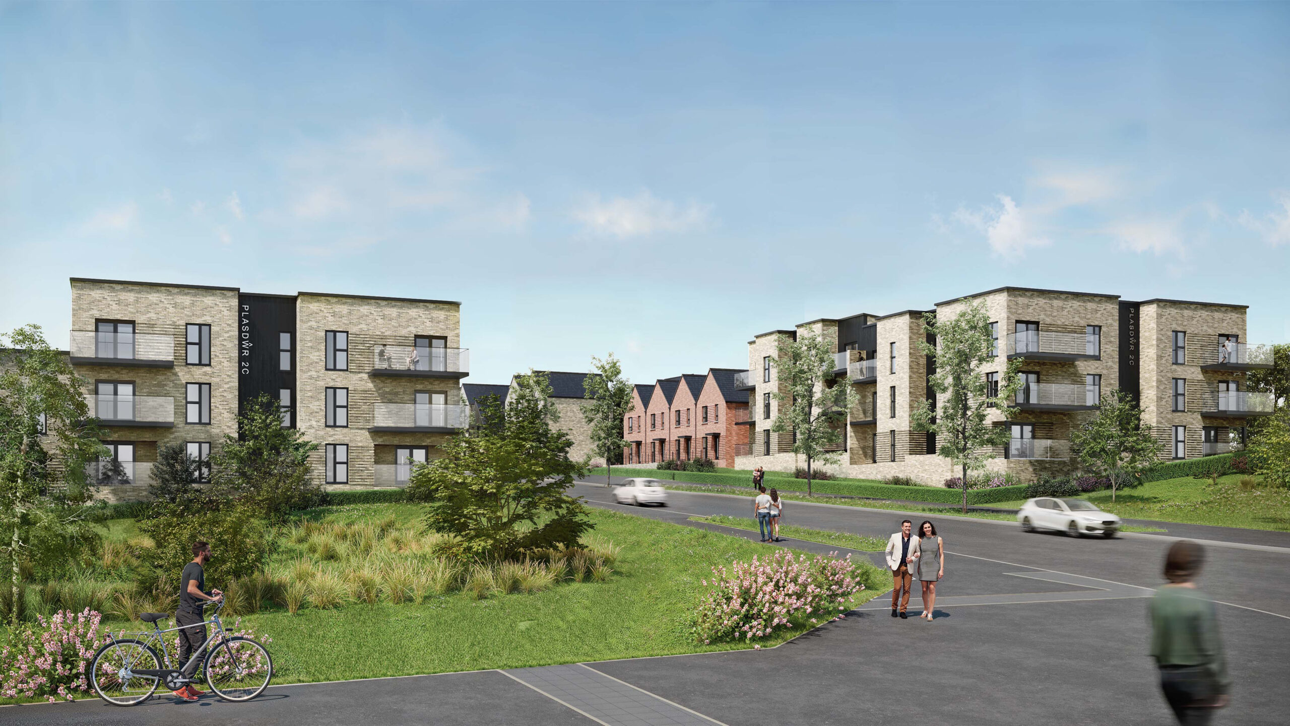 A long shot of a housing estate showing two blocks of flats and a row of houses, with a grass verge and people walking on the pathway, two cars driving past and one person pushing a bike