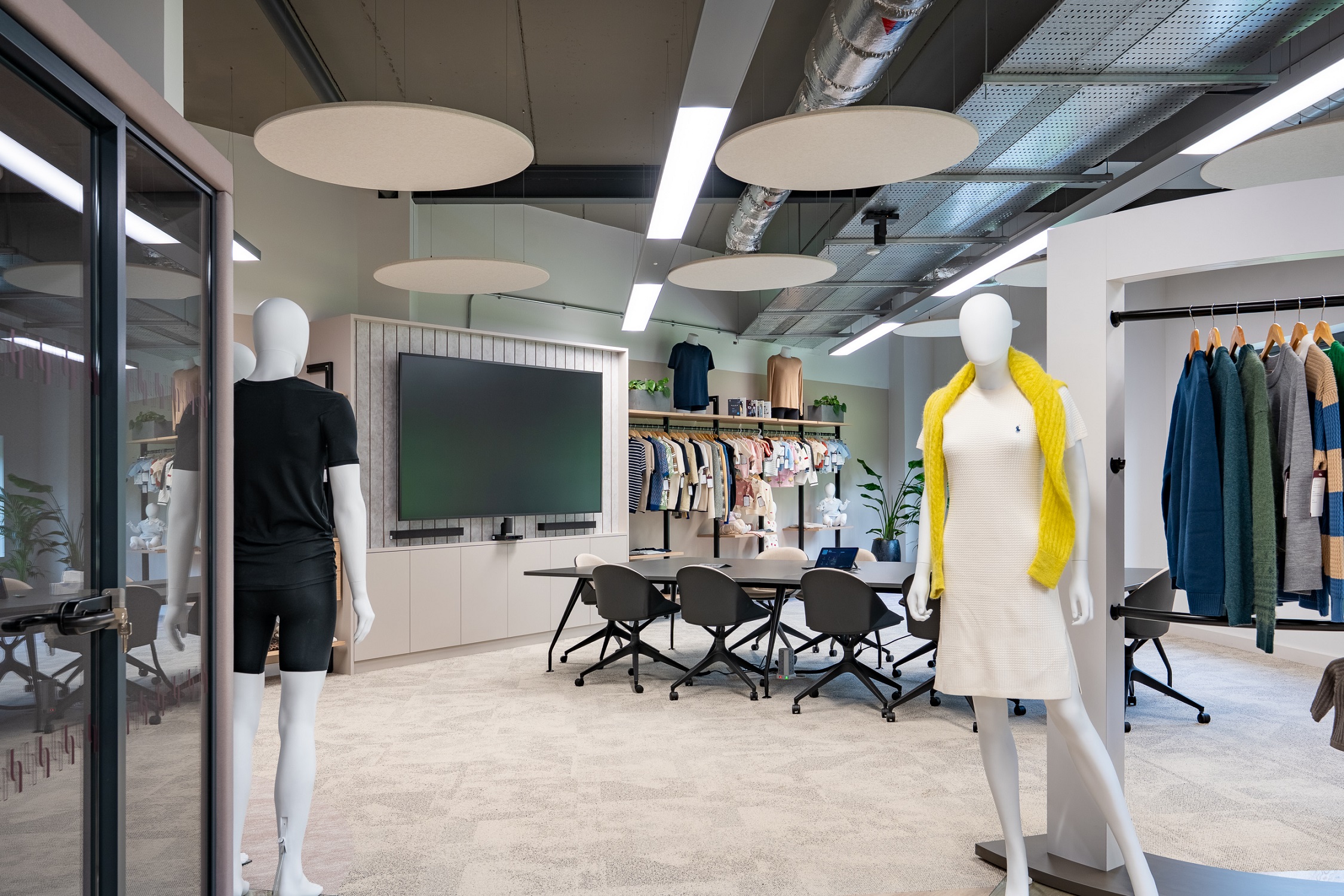 A spacious room with 2 mannequins placed sparingly around the room, displaying a black t-shirt and a yellow scarf with a clothes rack with jumpers and a meeting desk with chairs and a corresponding television in the middle of the room