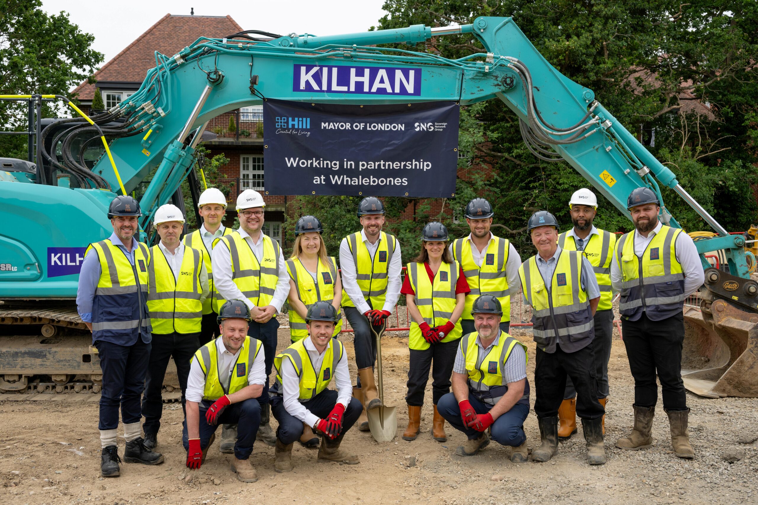 The Hill Group team wearing hi-vis vests and workwear stood together for a group photo on a building site with a turquoise digger in the background and a promotional Hill Group banner