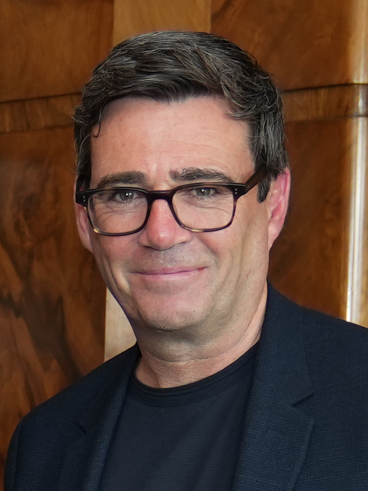 Headshot of Andy Burnham, the UK Prime Minister amongst a brown, wooden background