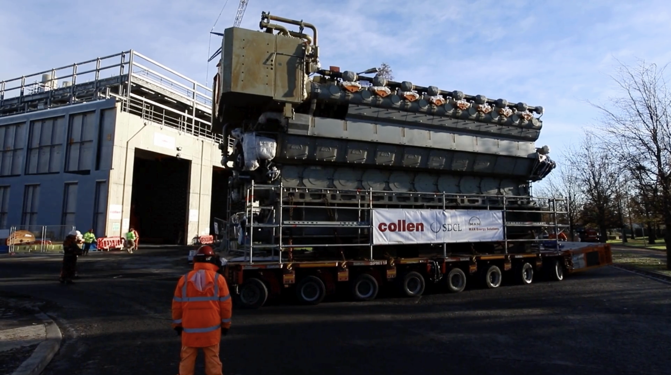 A man wearing an orange high-vis jacket and pants, wearing a red hard hat on a building site, looking at a big lorry with the sign 'Collen' on the front