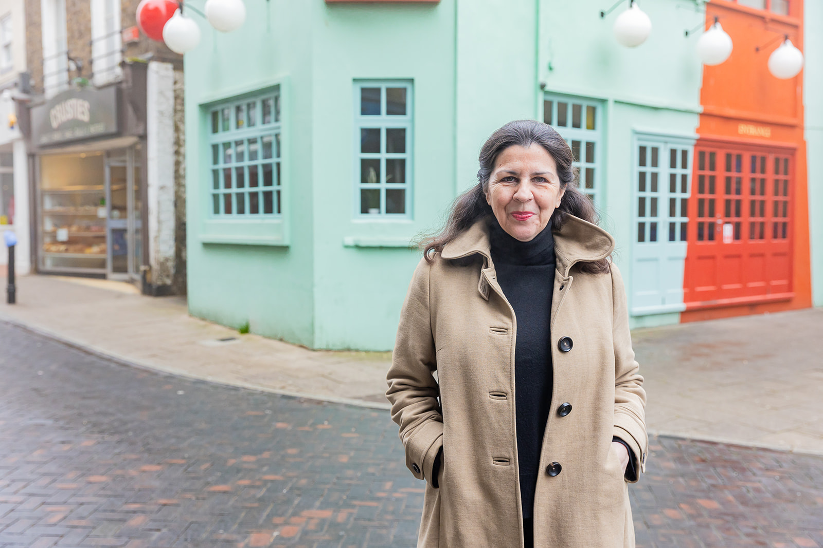 Emma Jones, the Small Business Commissioner, smiling and wearing a long nude coat with her hands in her pockets stood on a street on the pavement with a turquoise and dark orange in the background