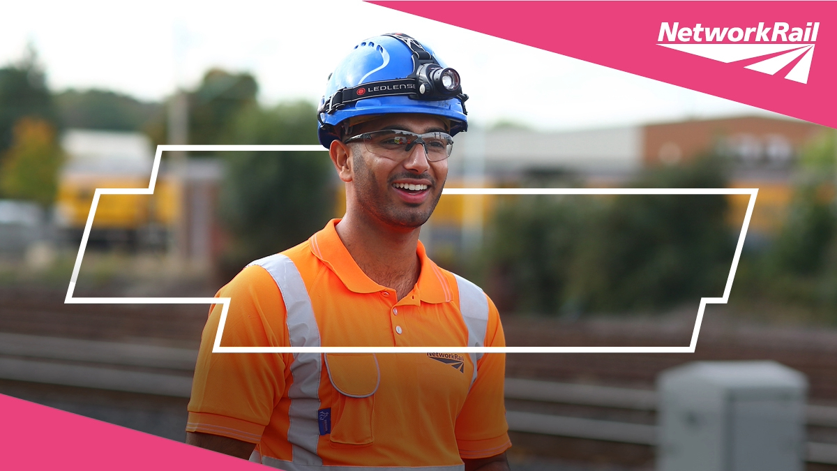 A man smiling in a blue hard hat in a high-vis, orange Network Rail t-shirt with a vivid background of a railway line