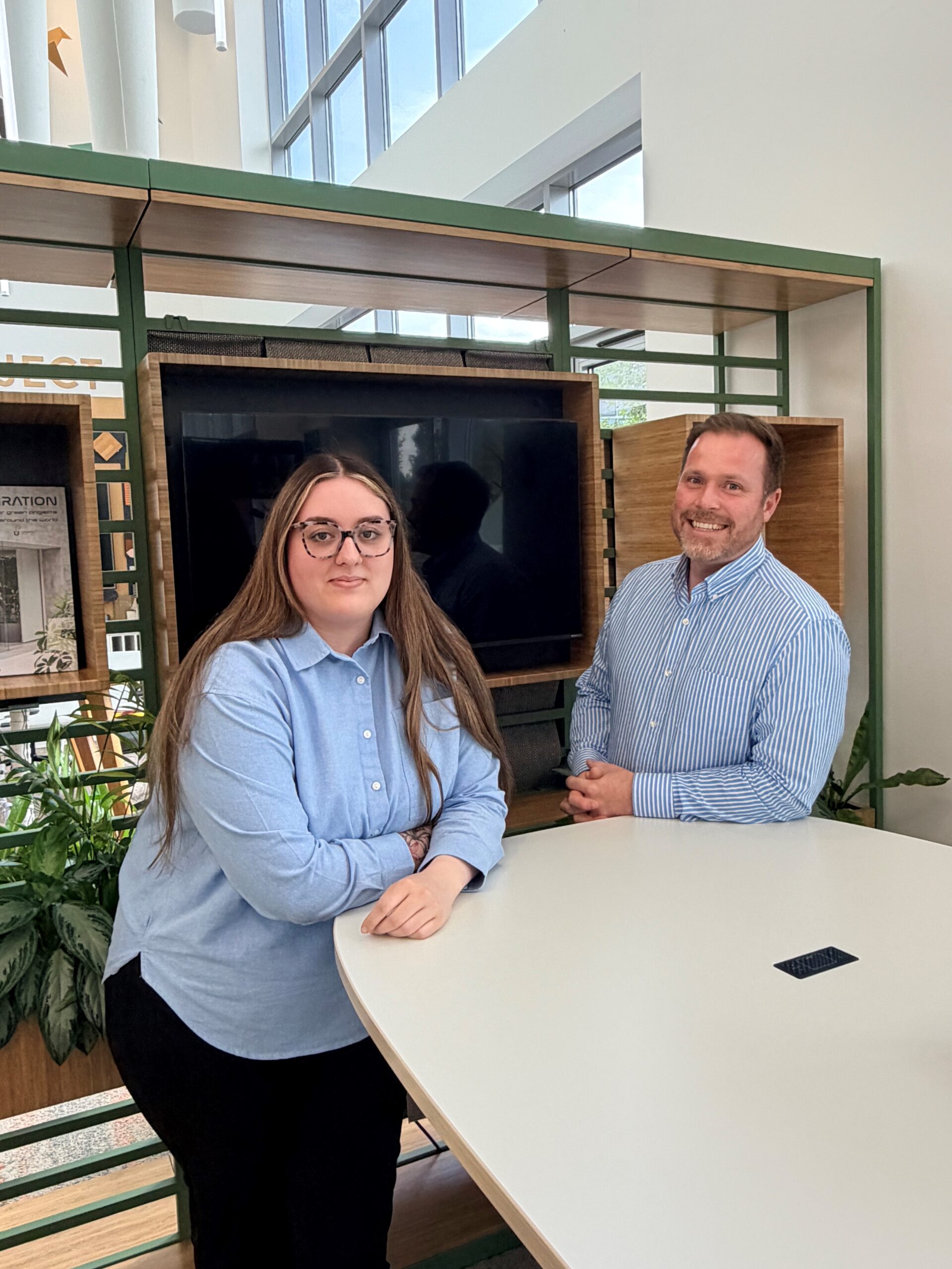Gemma Wort, Accounts Assistant Apprentice and Tom Beeson, Associate Project Director of Blueprint Interiors, leaning over a grey table with a shiny black wall, and an office backdrop, in the background