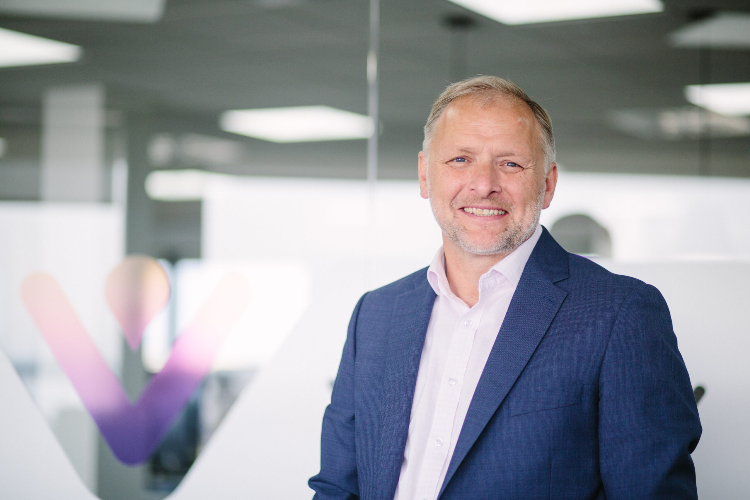 A headshot of Mark Perry, Chief Executive of VIVID, smiling wearing a white shirt and a navy blazer