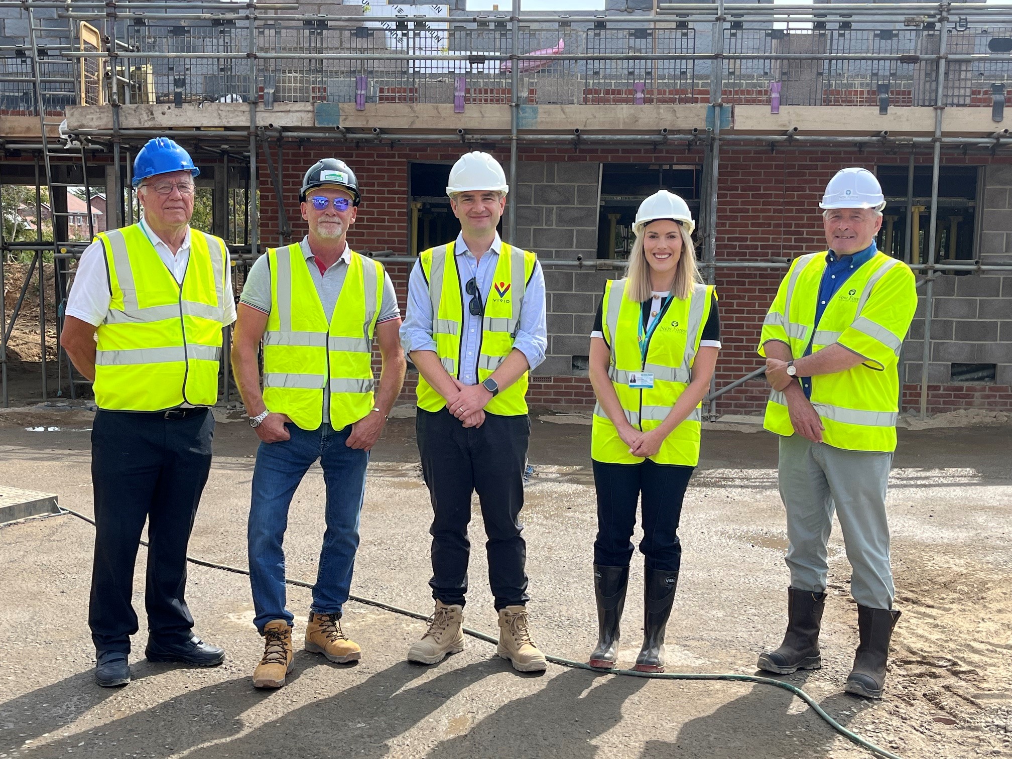 4 men and 1 women stood in a line wearing high-vis jackets & hard hats, stood on a building site, stood in front of a new house which is in the progress of being built, supported by scaffolding