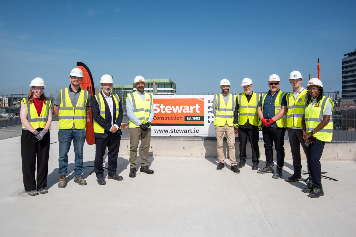 A team of builders wearing high-vis jackets and white hard hats, stood either side of the 'Stewart Construction' which is hung on a fence