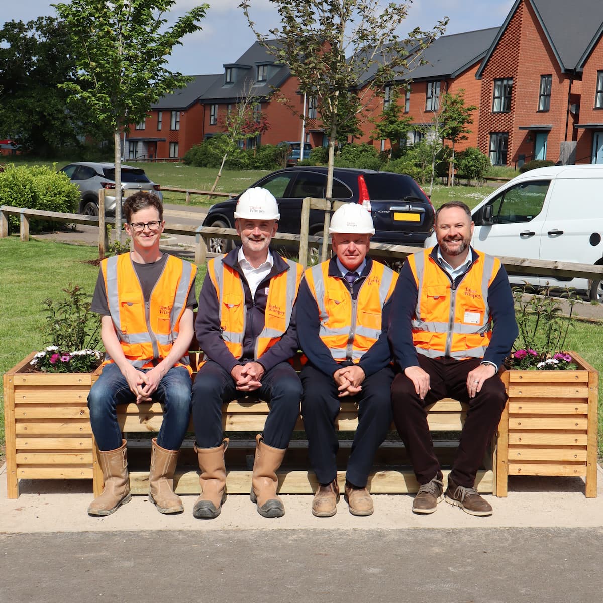 Four work men in orange hi-vis vests sat on a garden bench in front of a grass verge with houses in the background