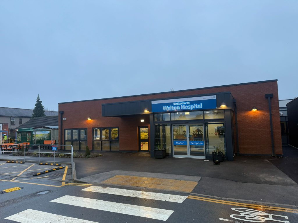 A shot of the front exterior, showing the entrance of NHS Walton Hospital, across the road from a zebra crossing and a cycle lane