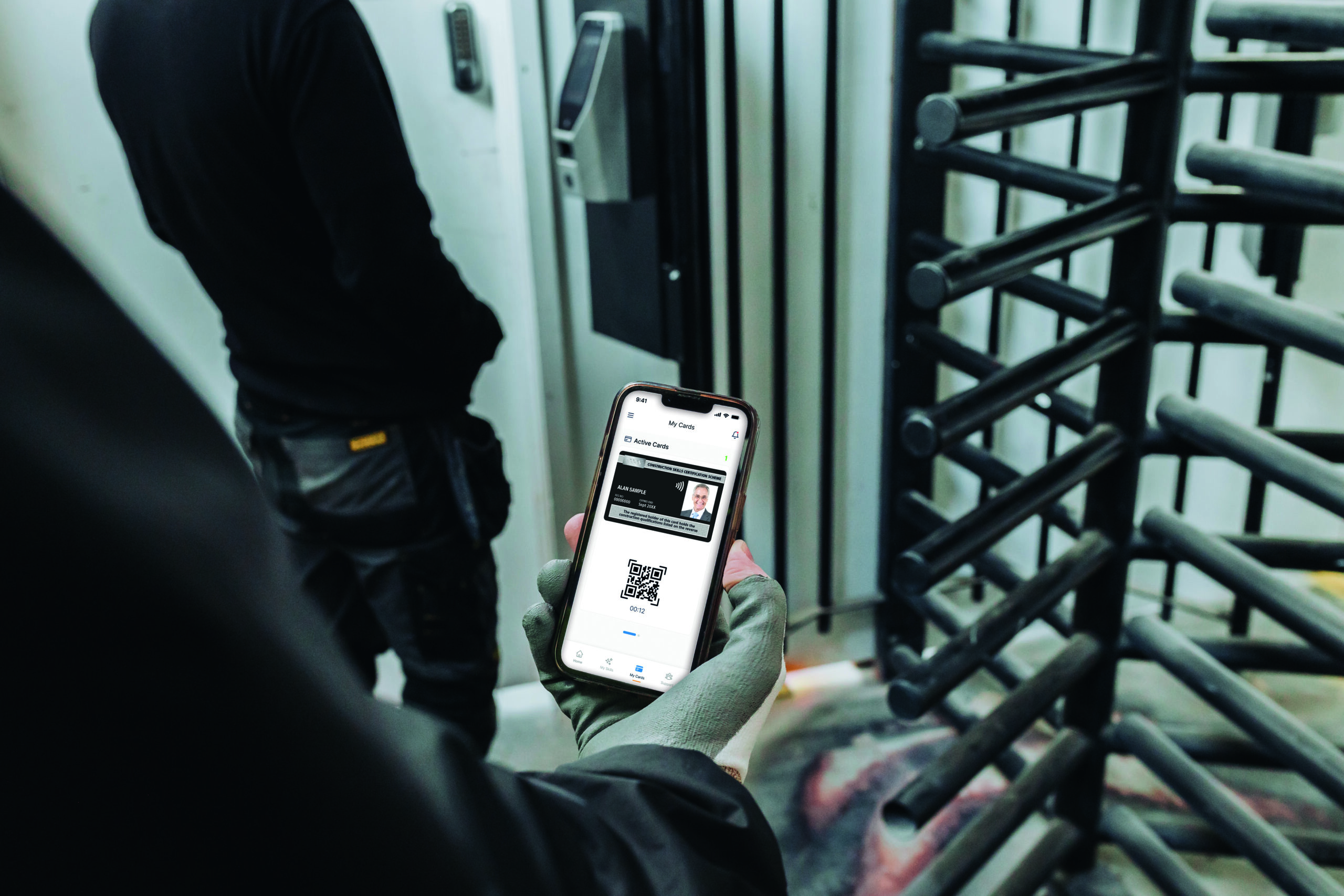 A man shown from behind wearing all black, with a person behind him wearing a grey glove, holding a phone, showing an ID card with a QR code, about to enter a locked entrance with security bars
