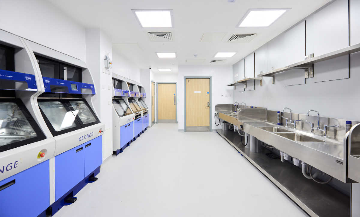 A long room with four silver sinks one one side of the wall and 6 blue machines with screens on the other
