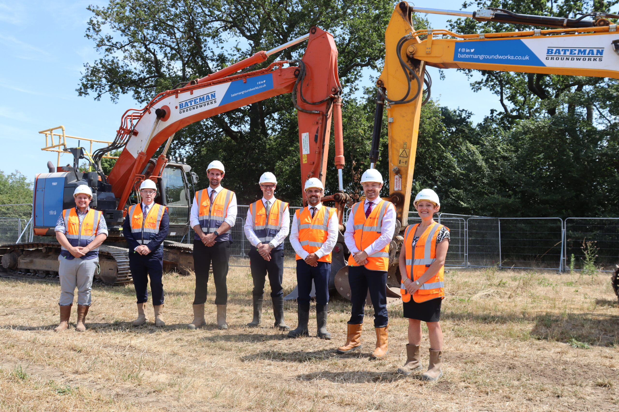 home-staging_01 Seven workmen stood in orange high-vis jackets, wearing hard hats on a construction site, in front of a digger