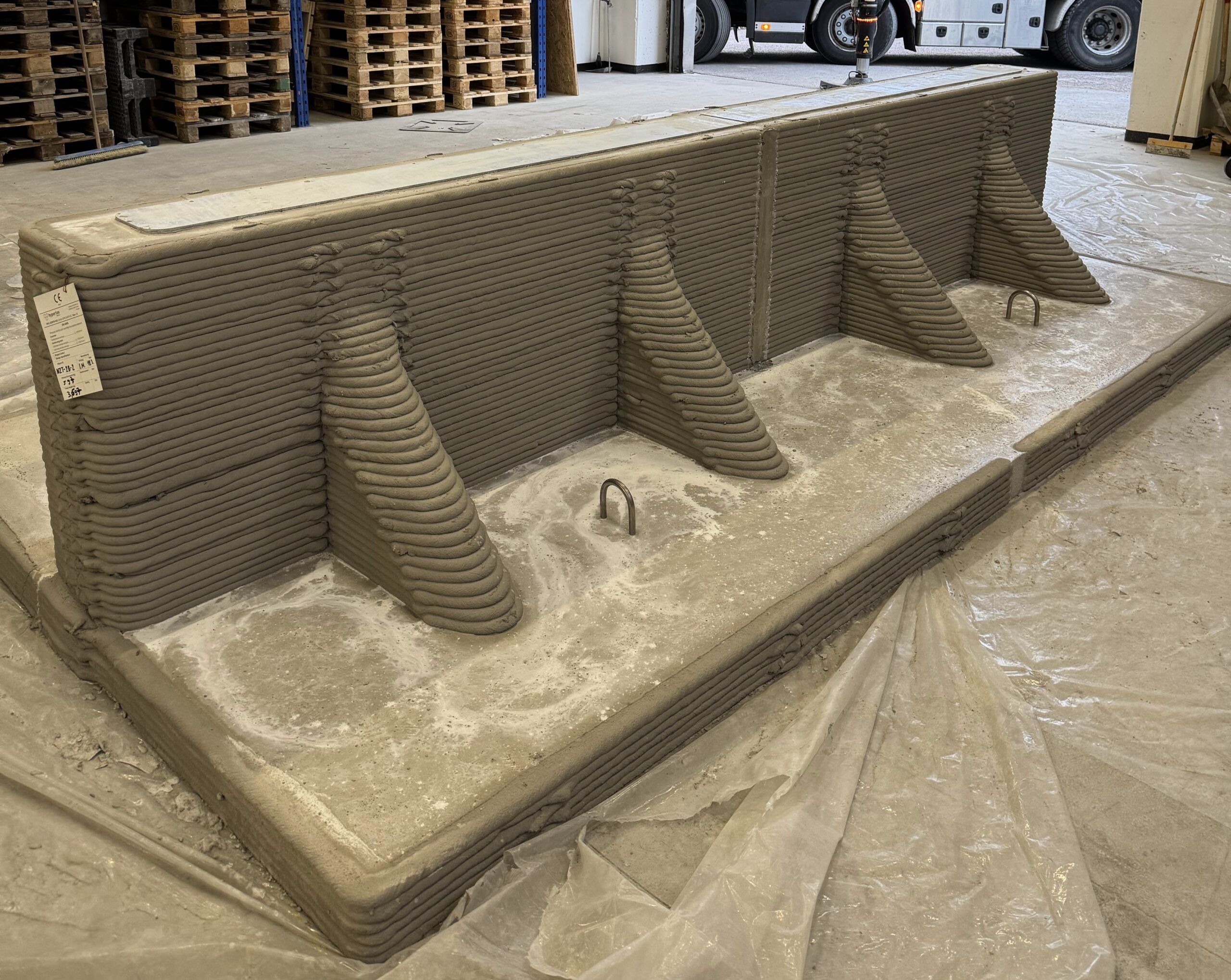 3D printed brown concrete sleepers in a vacant building and the shutters partially open with a workman in orange high-vis clothes in the doorway