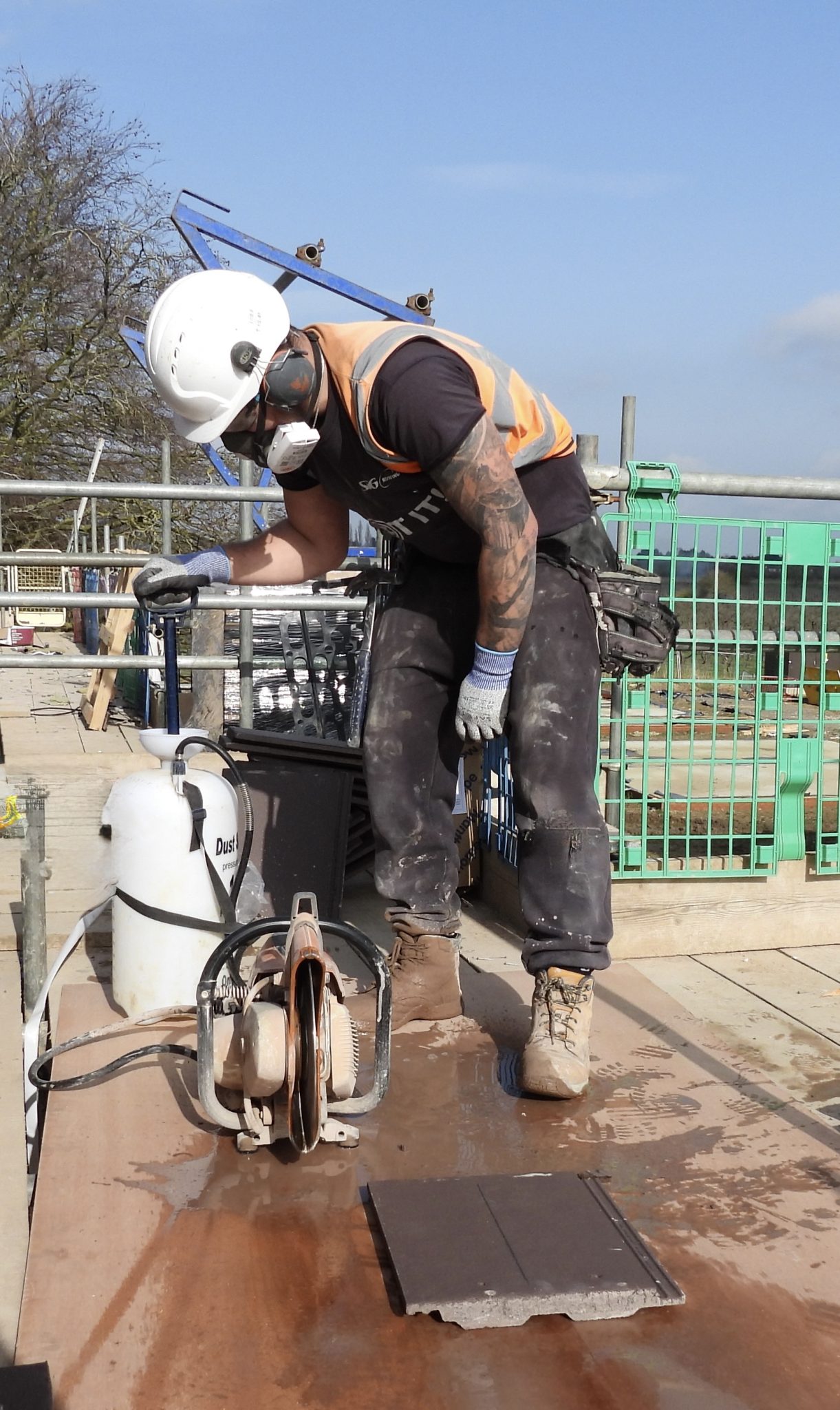 A workman stood working with equipment on a dirty building site, wearing a white hard hat, a protective mask and an orange high-vis jacket