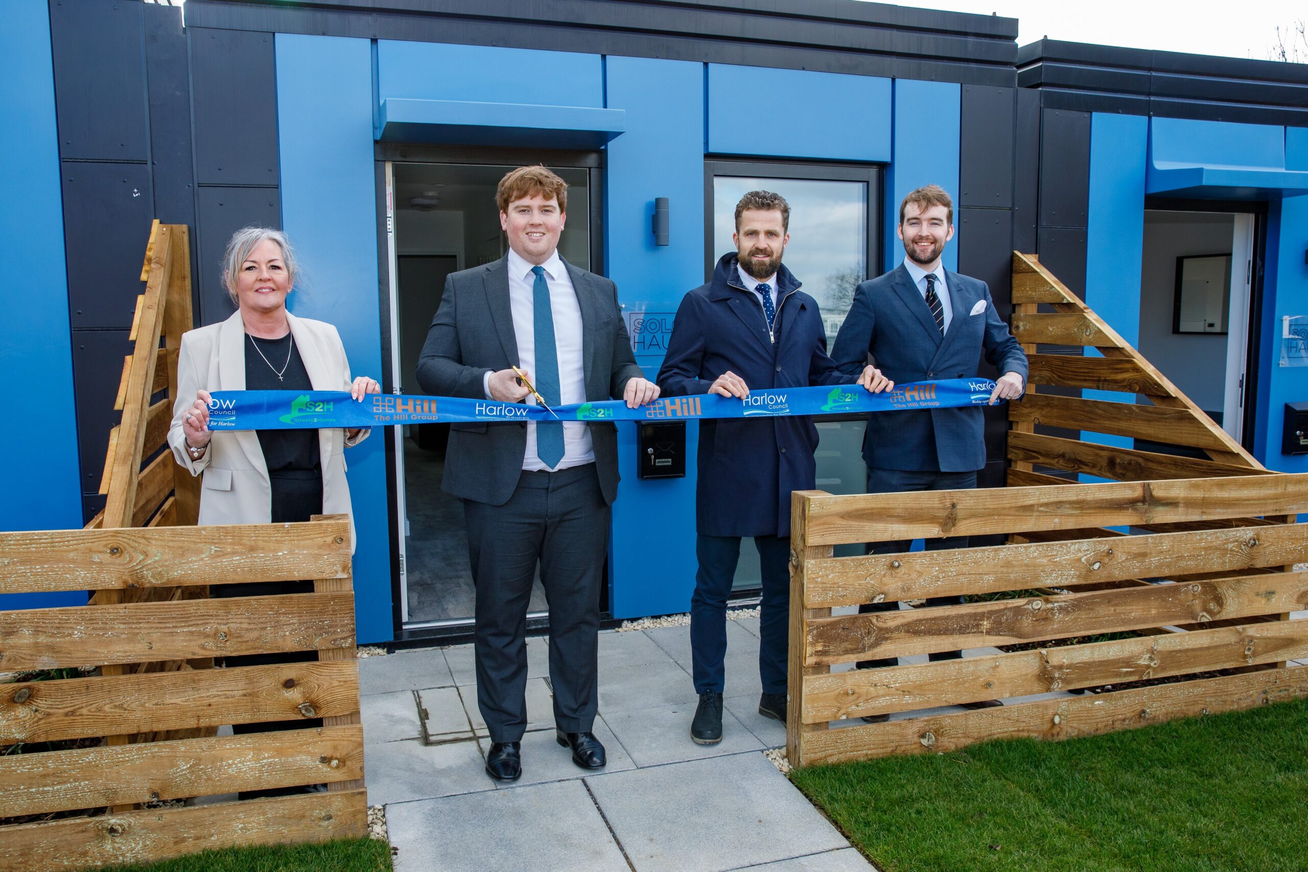 One woman and three men stood outside a small blue building, being stood behind blue ribbon tape