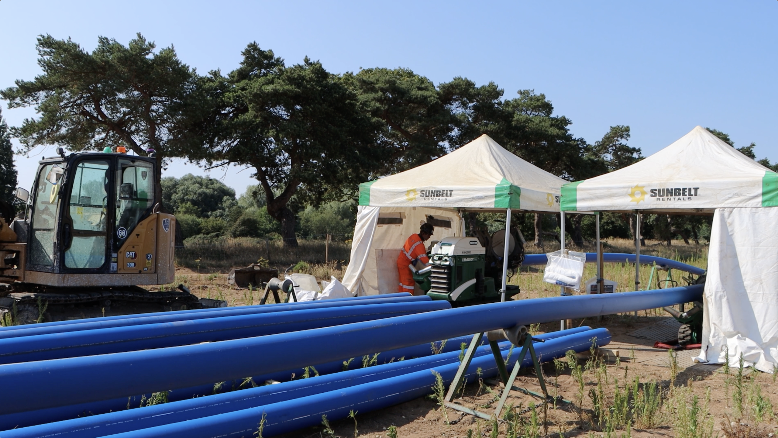 Stoke Ferry showing a blue Pipeline and a mini digger in a field surrounded by trees and 2 gazebos