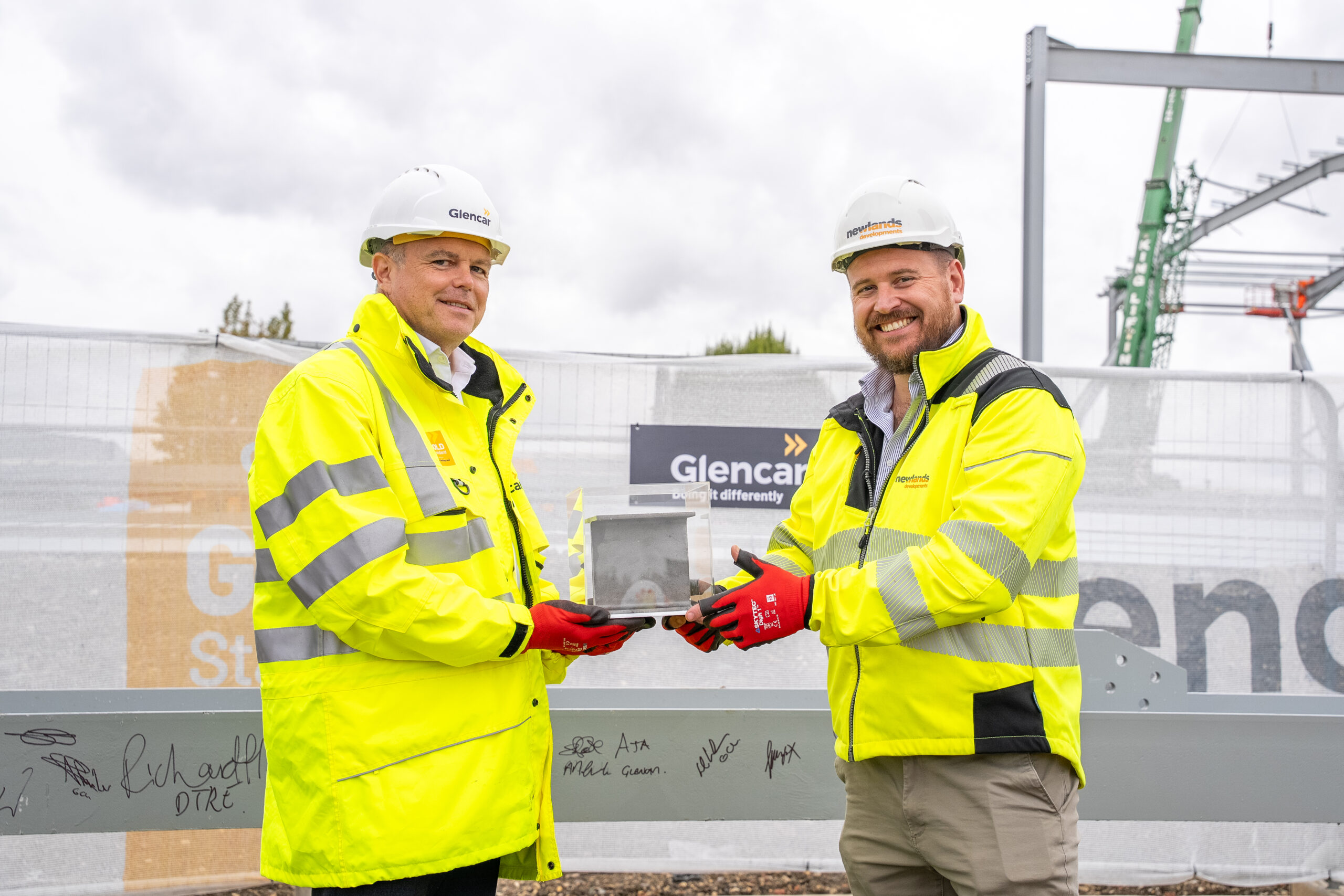 Two construction professionals in high-visibility clothing and hard hats hold a signed steel component at a building site, with construction equipment and Glencar and Newlands Developments branding visible in the background.