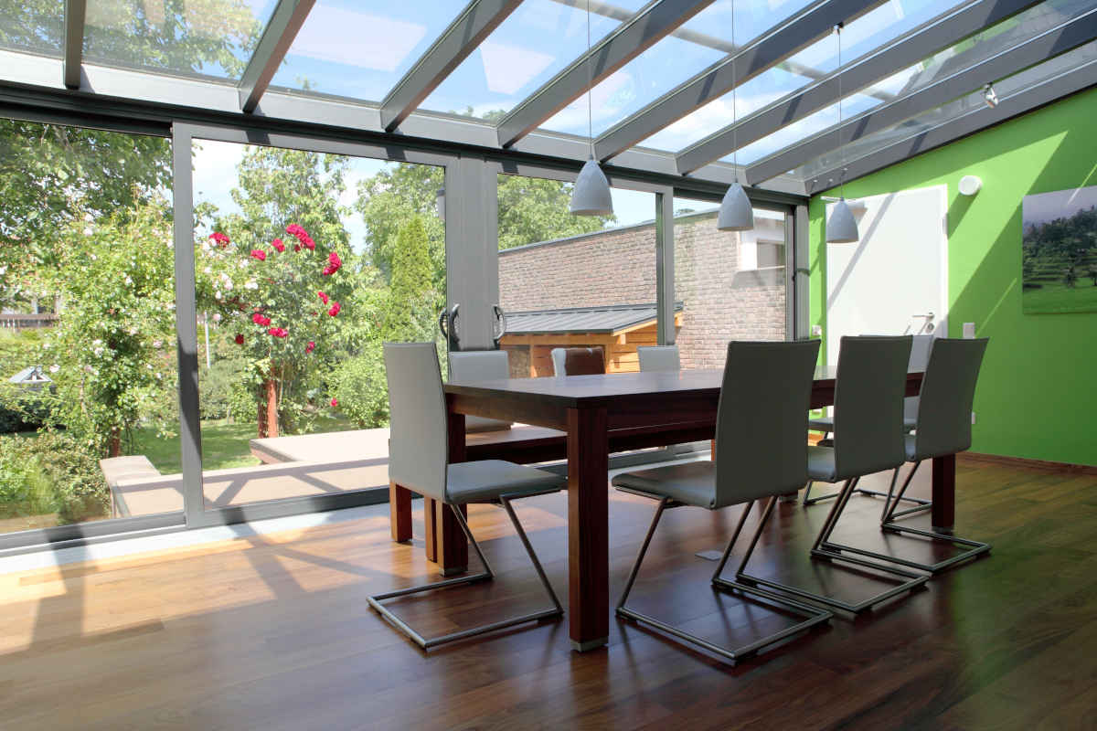 A wooden table surrounded by eight cream chairs in the middle of a room with a glass ceiling and windows looking onto greenery