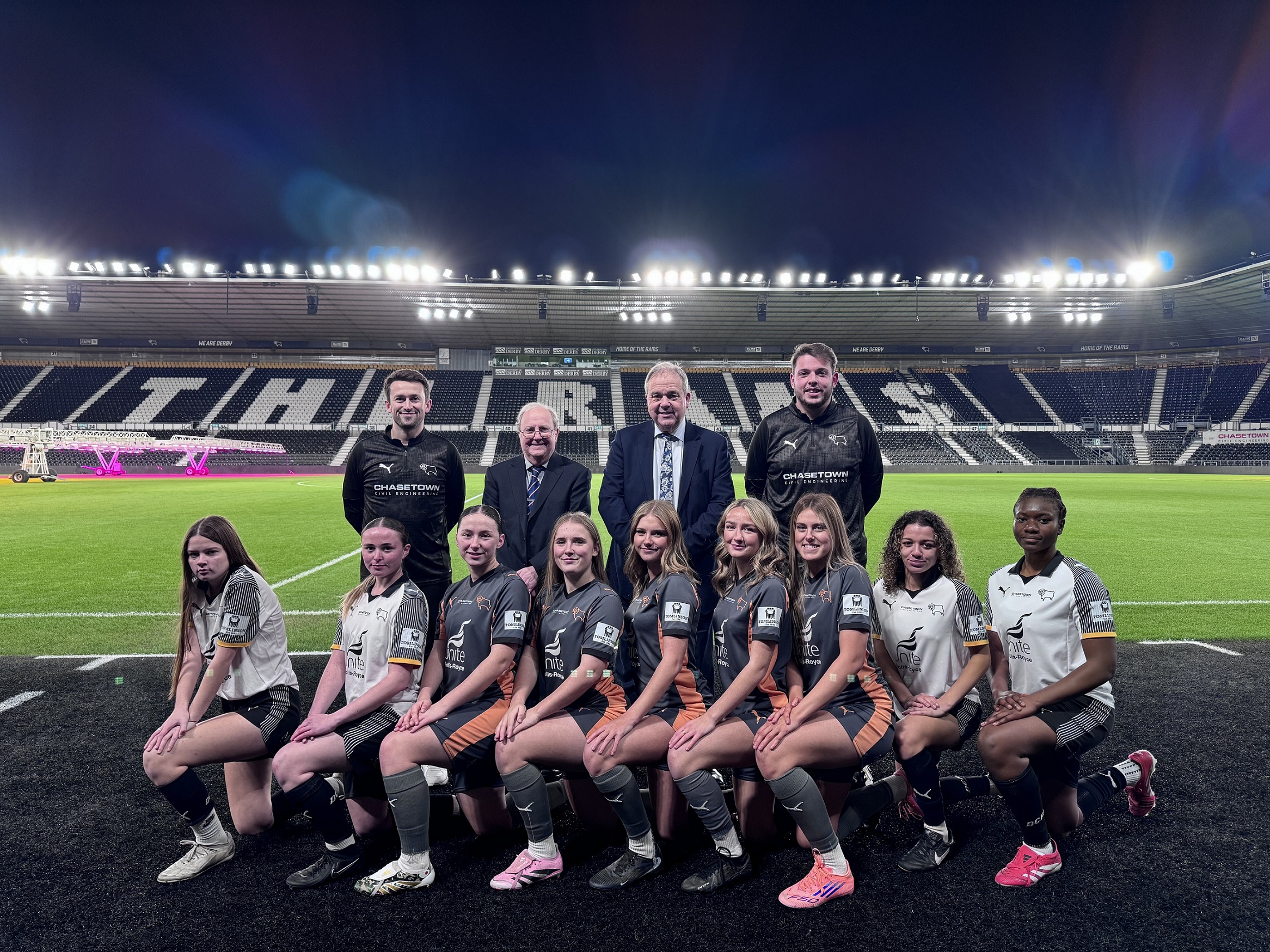 home-staging_01 A team of nine girls in their football kit, crouched in front of training staff and two men in a suit and tie on the touchline of a football ground