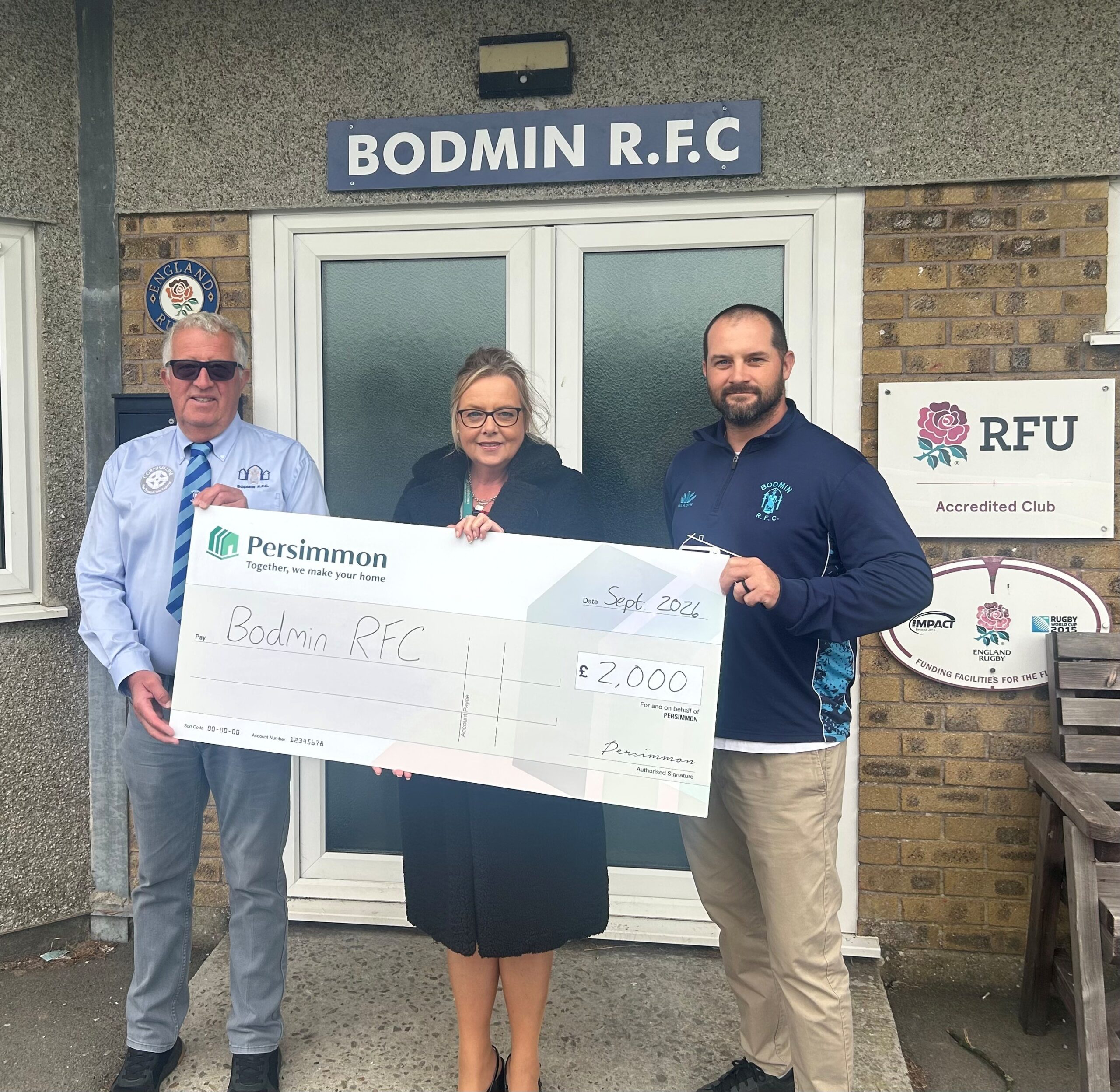 A woman stood between two men with them all holding a large cardboard cheque from Persimmon indicating: 'Bodmin RFC' - £2,000'