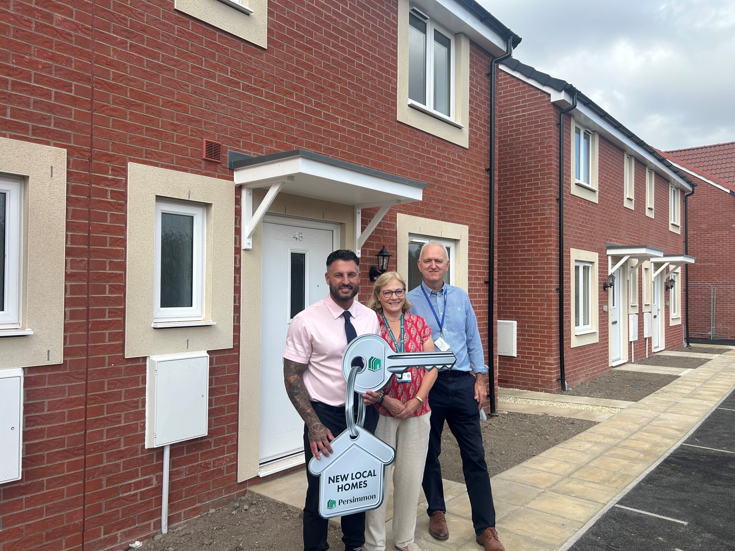 A woman stood in between two smartly dressed men, with the man on the left holding a large cardboard cut out of a keyring stood outside of a semi-detached house