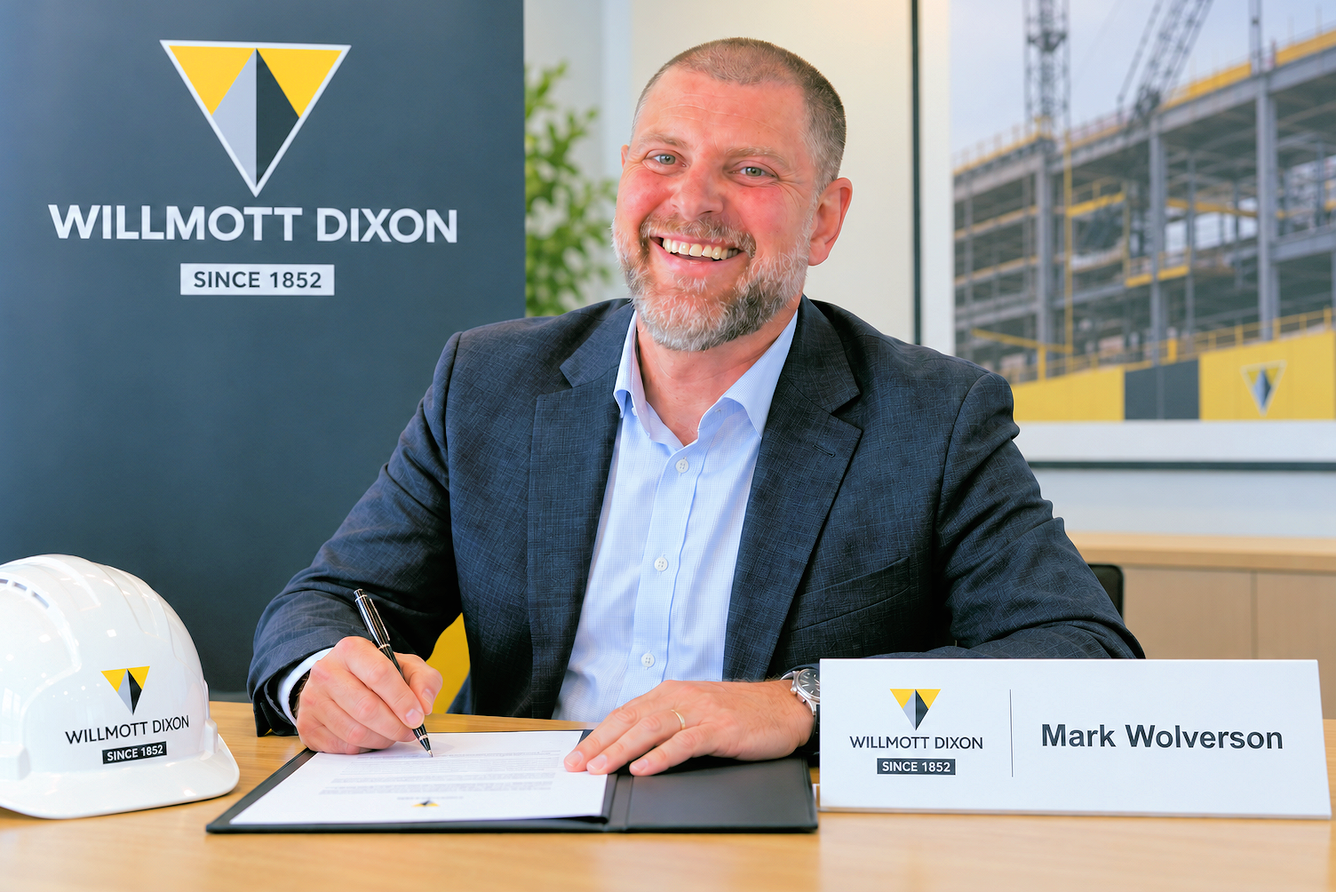 Mark Wolverson, North East Delivery Director at Willmott Dixon, signs a document at a desk, with Willmott Dixon branding, a hard hat and construction project imagery visible in the background.