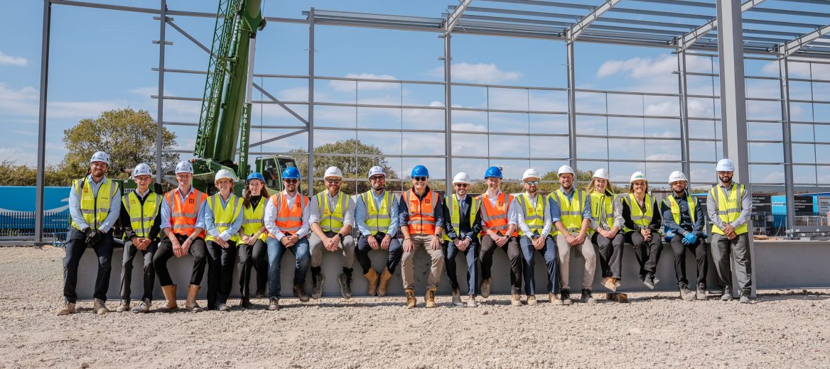 Seventeen work people wearing high-vis jackets and hard hats amongst scaffolding and empty land