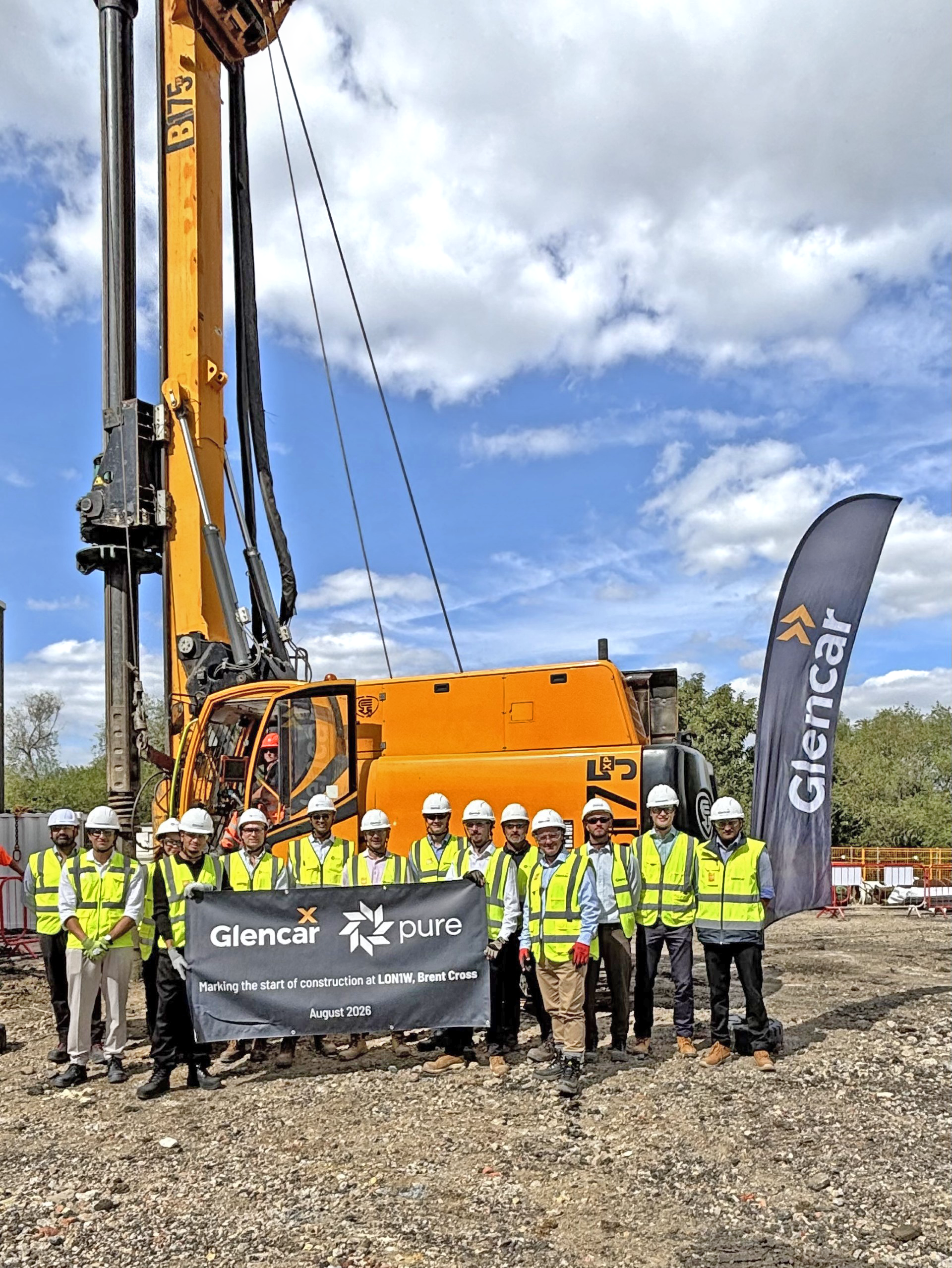 Pure DC & Glencar Stakeholders stood in a line holding a sign, wearing hard hats and high-vis jackets and in front of a big orange digger