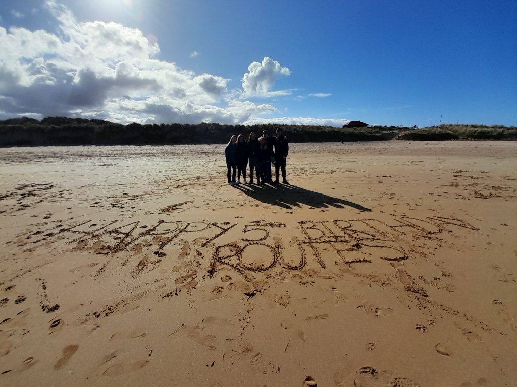 Five people stood on a beach with: 'Happy 5th Birthday Routes' carved out in the sand