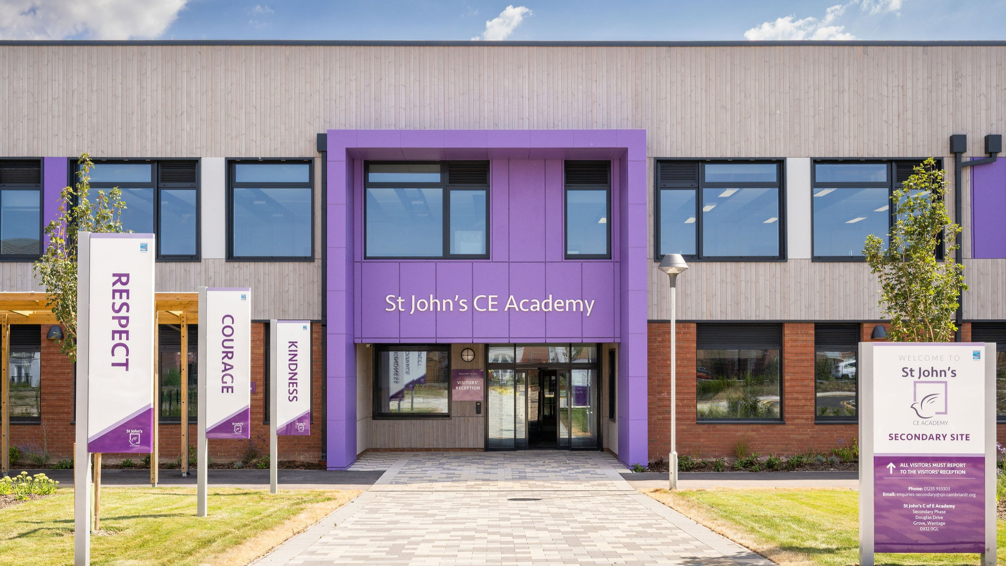 The front of St John's CE Academy, with a grey path and a row of pols with purple flags along the grass verge leading to the entrance