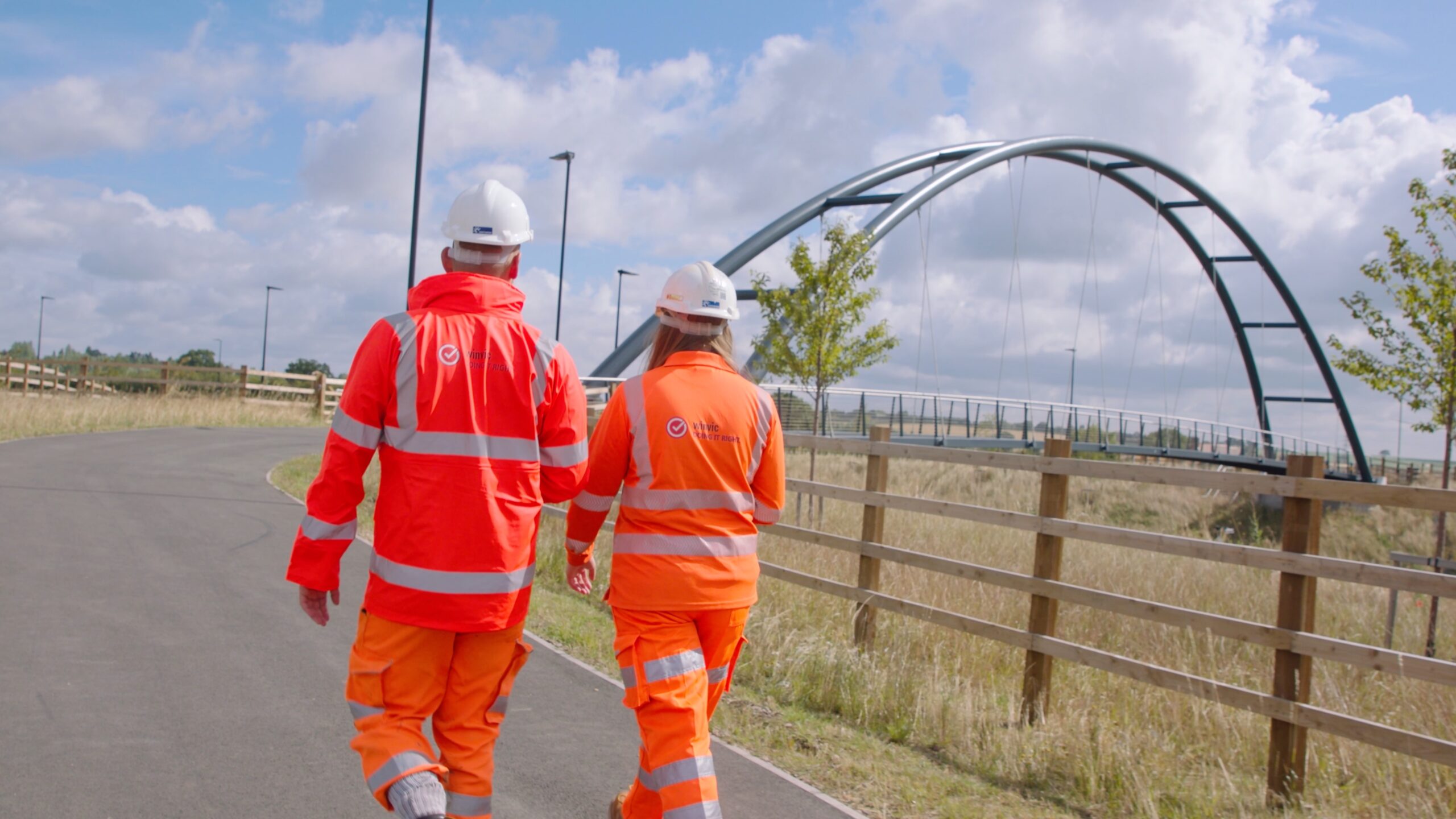 The back of one woman and one man wearing orange high-vis jackets, pants and white hard hats, walking down a path which is surrounded by long grass