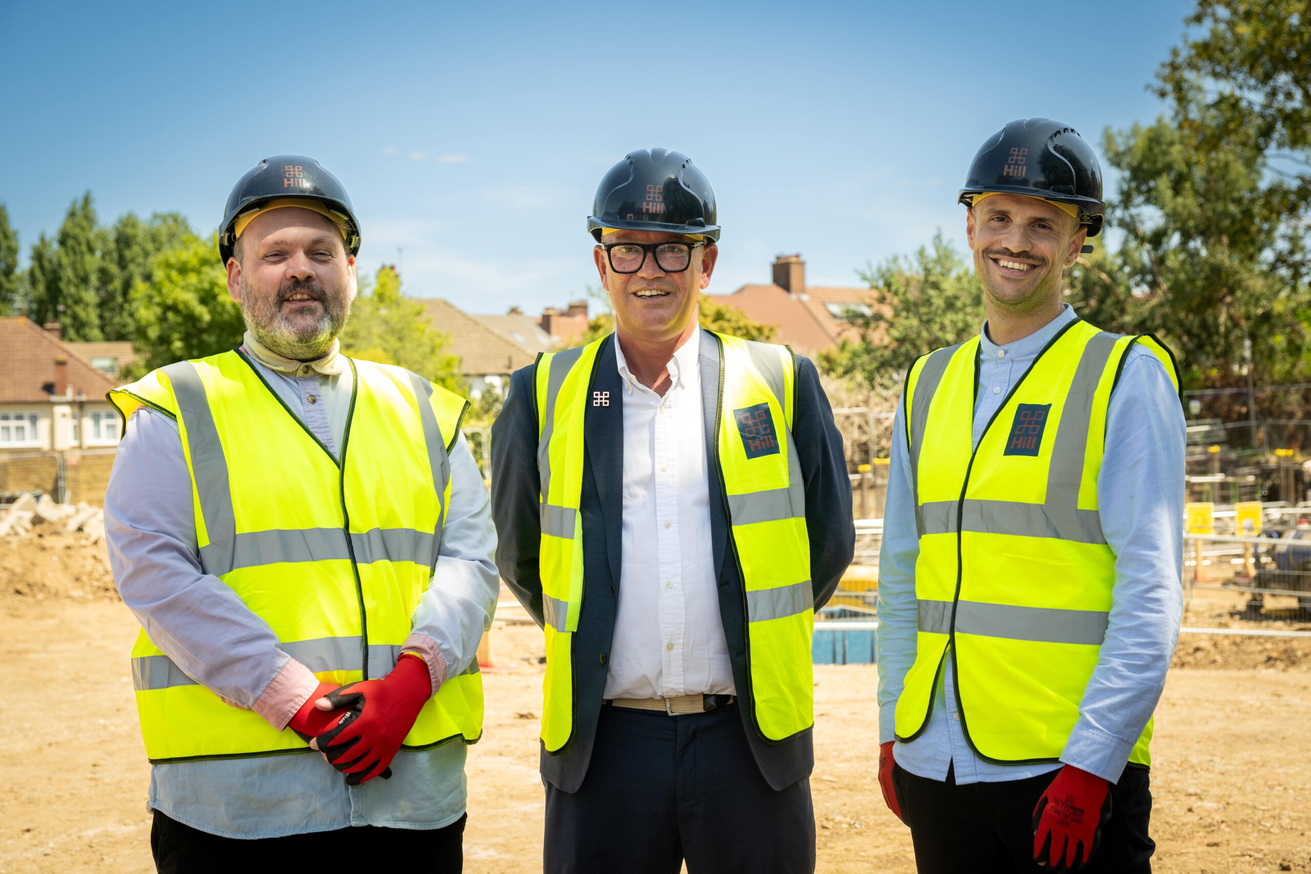 Three men dressed in a shirt and pants, wearing high-vis jackets, red gloves and Hill hard hats on a building site