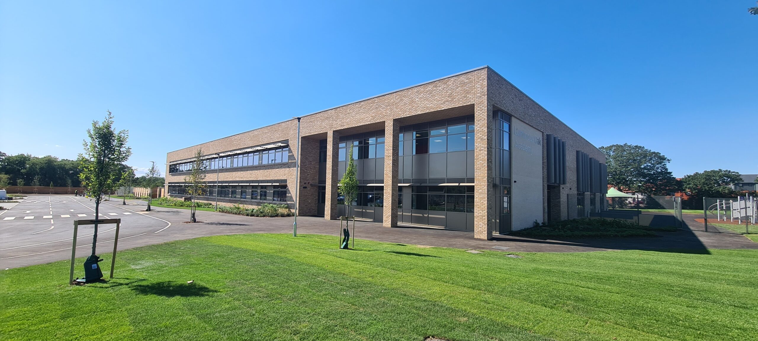 A brown building with a silver fenced play area to the right and a grass verge with a car park