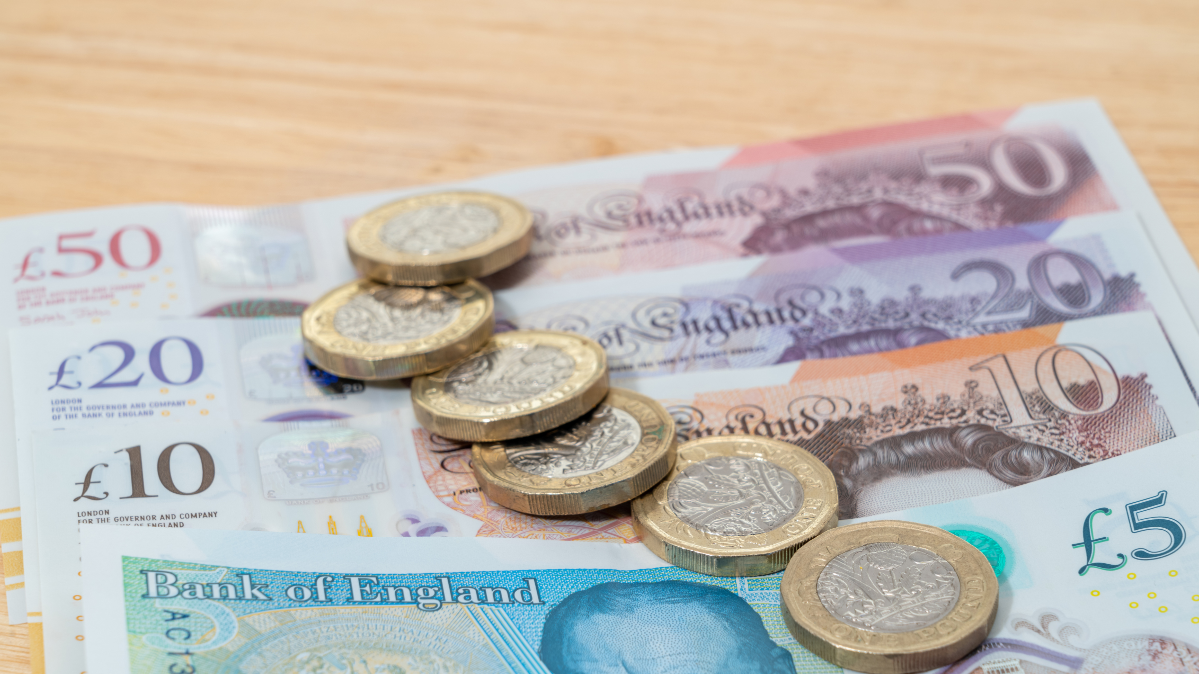 A fifty pound, twenty pound, ten pound and five pound note on a wooden table with six one pound coins on top