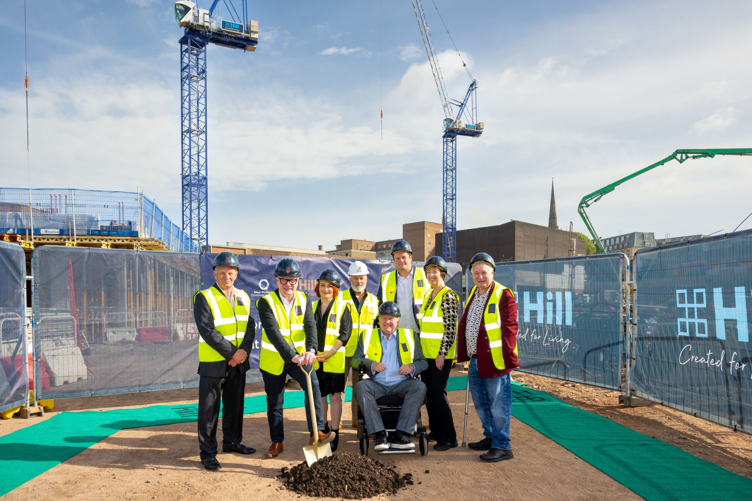 Seven older people wearing high-vis jackets and hard hats, with one in a wheelchair stood on a building site with a shovel in front of Hill-branded fencing
